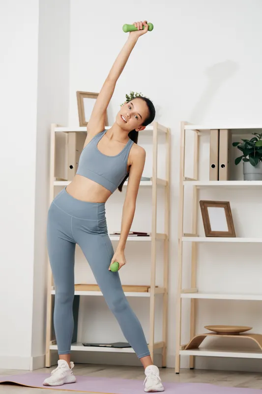 Fitness woman exercising at home with dumbbells, stretching to the side. Modern athletic wear, soft lighting, minimalist shelves, and plant accents create a calm workout scene.
