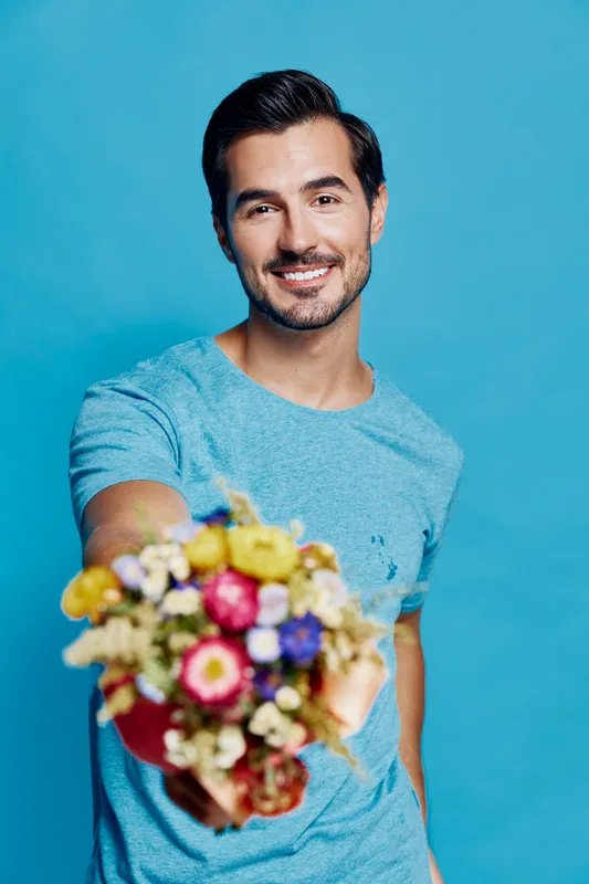 Happy smiling man holding a colorful bouquet of flowers and looking at the camera against a solid blue background. Bright, cheerful mood concept.