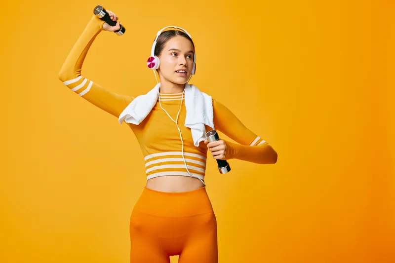 Fit young woman in vibrant orange activewear lifts dumbbells with headphones and towel in a studio, conveying energetic fitness, strength training and confident workout lifestyle.