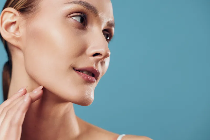 A closeup portrait of a woman with clear skin and a soft glow, touching her jaw with a calm expression against a blue background. Concept for skincare, beauty routines, and self care.
