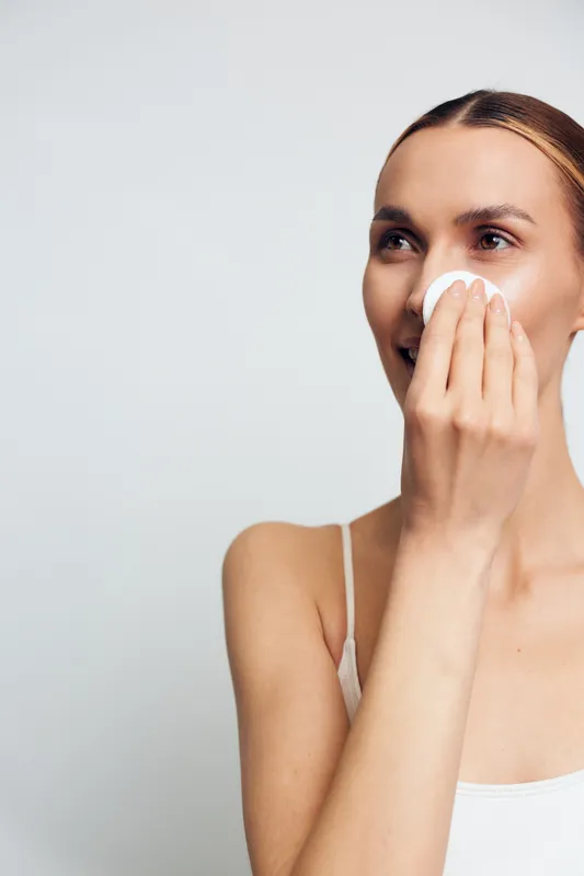 Young woman with clean skin removes makeup with cotton pad looking away smiling wearing white tank top isolated on light background skincare beauty routine concept.