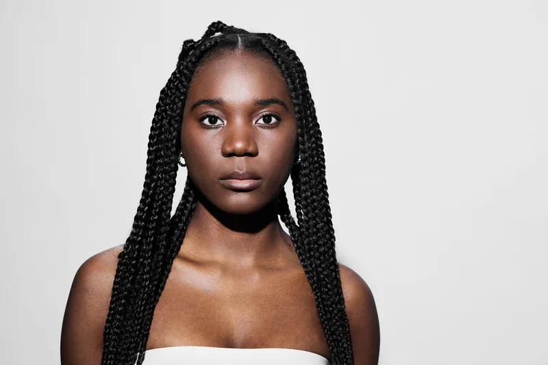 Portrait of a young Black woman with braided hair, wearing a simple white top, showcasing a serious expression against a minimalistic white background