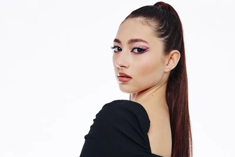 Young woman with sleek dark ponytail and artistic pink eye makeup gazes confidently over her shoulder. Studio portrait on white background. Fashion and beauty creative concept.