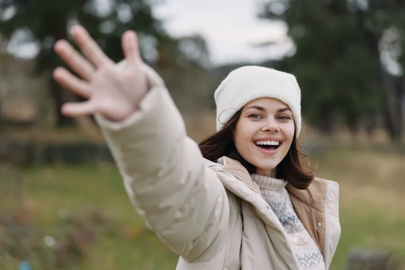 Happy woman outdoor reaching hand, joyful expression, wearing winter coat and beanie in an autumn park, creating a candid portrait with friendly gesture and natural mood.