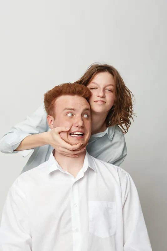 Playful young woman with curly hair teasing redhead man in white shirt on light background. Concept of friendship, fun, humor, and casual lifestyle in studio portrait.