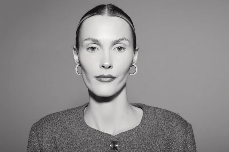Portrait of a fair skinned Caucasian woman with slicked back hair wearing hoop earrings and a textured jacket on grey background.