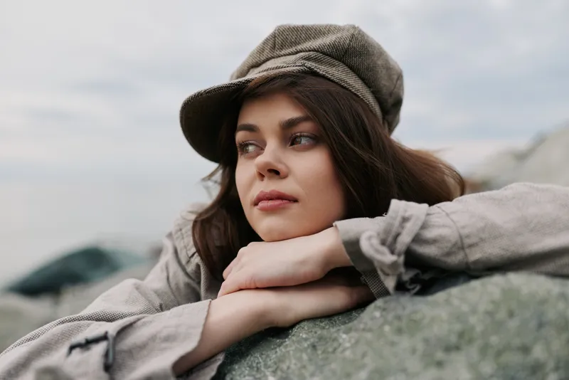 Young woman in casual clothes and cap leans on a rock outdoors. She looks thoughtful and relaxed with natural makeup, soft light, and a blurred background. Concept for leisure and contemplation.