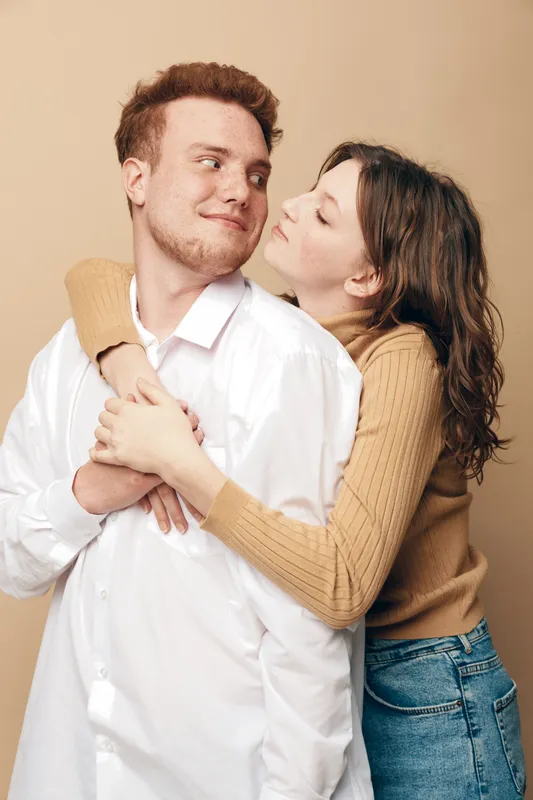 Young couple in casual clothes embraces tenderly against beige background. Woman hugs man from behind with affectionate smile. Concept of love, relationship, intimacy, and warmth.