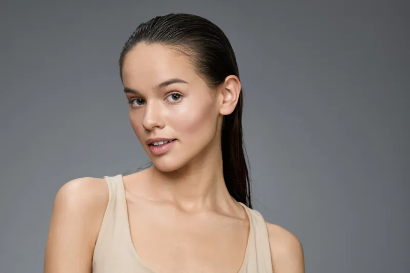 Young woman with wet hair and natural makeup, posing confidently against a gray background, showcasing healthy skin and a neutral outfit