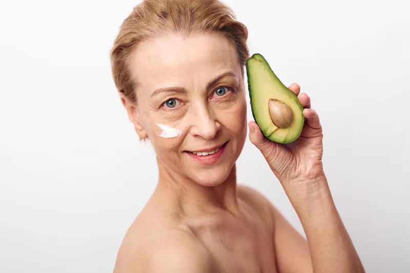 Mature woman with natural skin holds half avocado near face with cream on cheeks. Concept for skincare, healthy lifestyle, natural beauty, and anti aging care on white background.