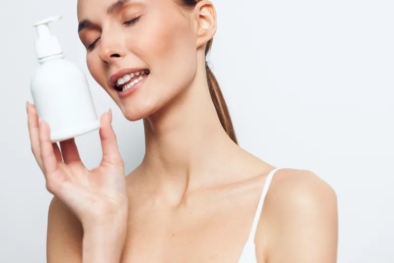 Young woman with clear skin holds white cosmetic bottle close to face with closed eyes and gentle smile isolated on white background, skincare and beauty concept.