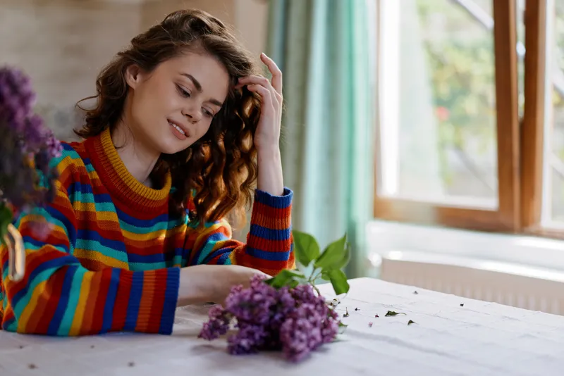 Smiling woman with curly hair sits at a table by a window, wearing a colorful striped sweater. Fresh flowers nearby create a warm, cozy home scene focused on lifestyle, comfort, selfcare, and natural