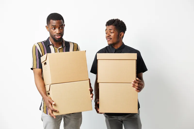 Two African American men carrying cardboard boxes, looking puzzled and surprised. Their expressions convey curiosity and confusion, lending a humorous touch to the scene.