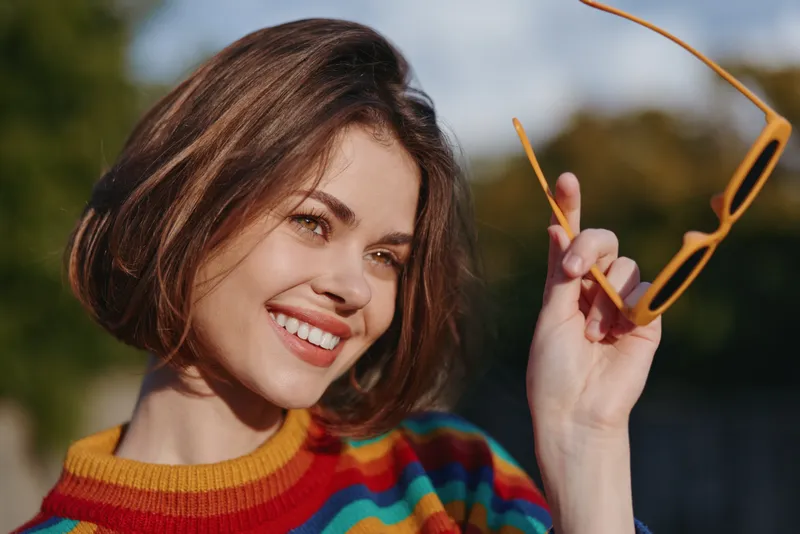 Woman, young adult with short bob hair smiling and holding yellow sunglasses, wearing colorful striped sweater in sunny outdoor portrait. Lifestyle vibe.