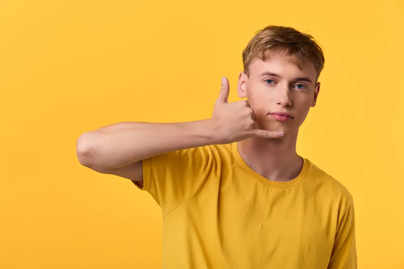 Hand gesture of a young man in a yellow shirt making a call me sign with thumb and pinky extended against a bright background, casual modern vibe suitable for lifestyle and social media use