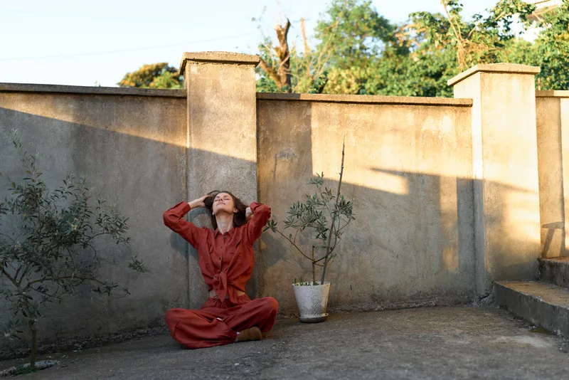 Joyful woman outdoors, seated in a red dress, eyes closed, hands overhead, relaxed pose, warm sunlight, soft breeze, rustic wall backdrop, potted plant nearby