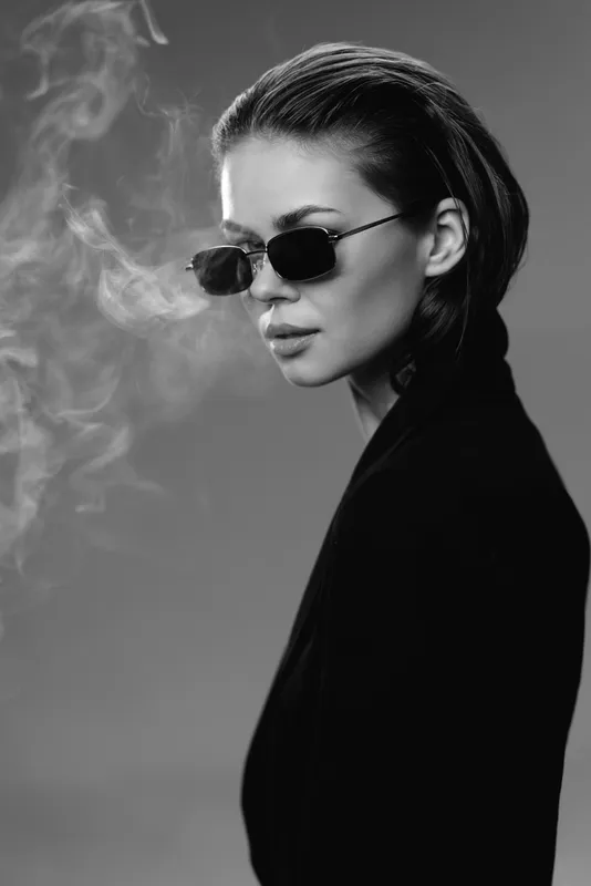 Black and white portrait of a stylish woman wearing sunglasses with smoky background and sleek hairstyle.