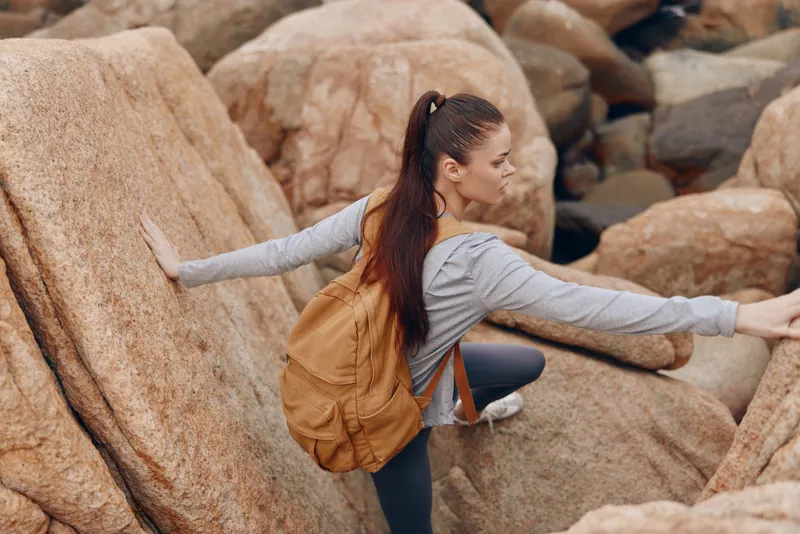Athletic young woman climbing rocky terrain, showcasing determination and strength in a natural outdoor setting with a tan backpack