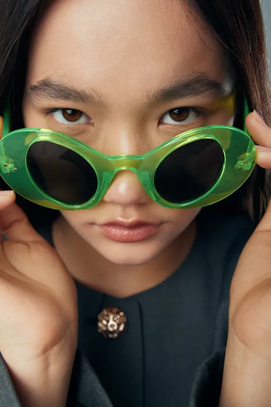Portrait of young woman with green sunglasses holding frames and looking at camera with intense gaze wearing dark jacket and brooch on front for fashion style closeup.