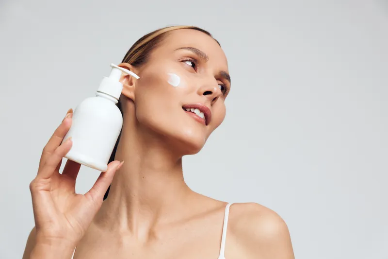 Woman applying face cream holding a white bottle near her cheek close up on a soft gray studio background concept of skincare wellness and self care gentle lighting smiling expression