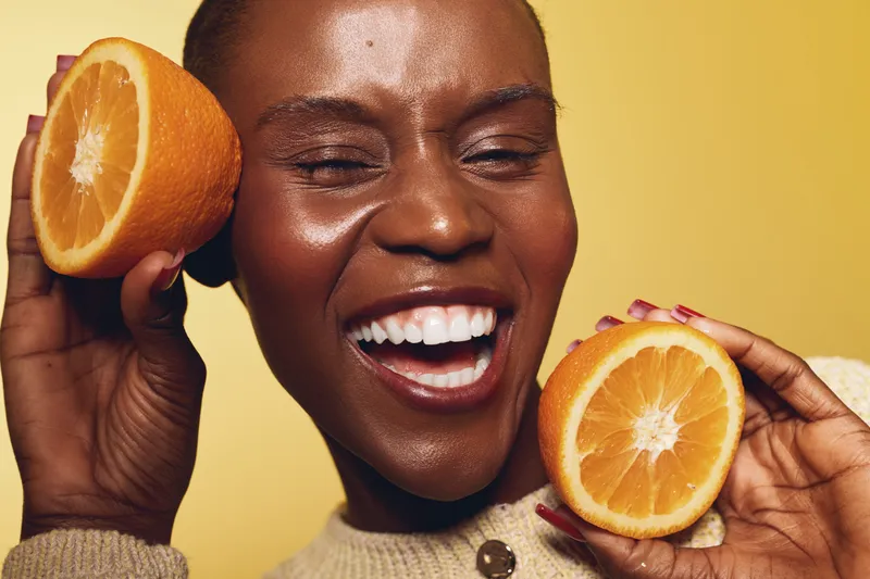 Happy African woman with dark skin holds two orange halves near her face, smiling widely against yellow background. Concept of fresh fruit, health, and natural beauty.
