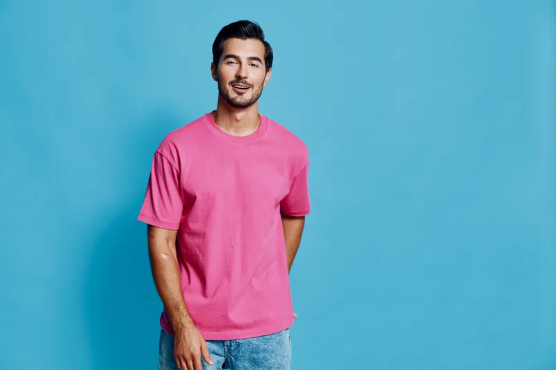 Full body cheerful smiling young man wearing pink t-shirt standing against a solid blue background, looking at the camera with a friendly expression. Fashion lifestyle concept.