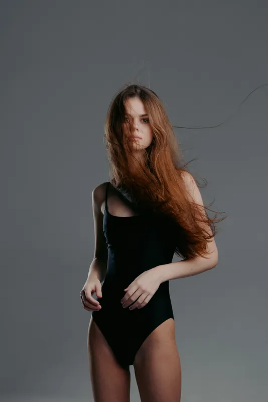 Young woman with long hair in a black bodysuit posing against a gray background with dramatic lighting and moody expression
