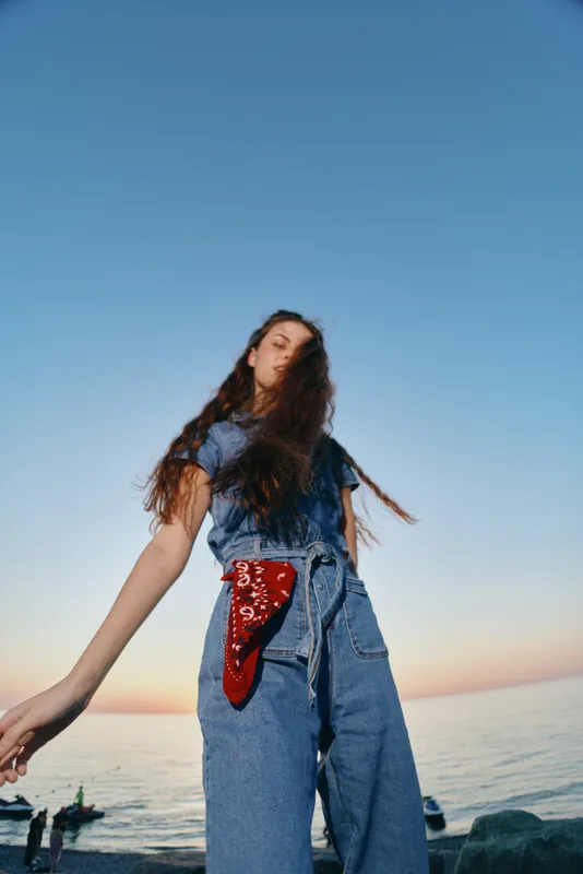 lifestyle woman wearing modern boho-western denim street style outfit with red bandana standing near ocean at sunset in film color tone