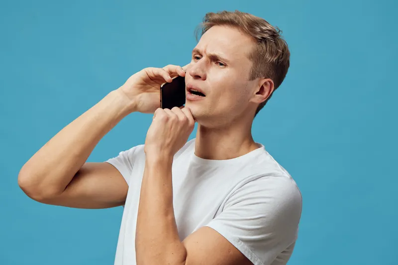 Modern young man with short hair making a concerned face while talking on a smartphone isolated on blue background. Studio portrait of adult male with expressive emotions, casual white t-shirt, and cl