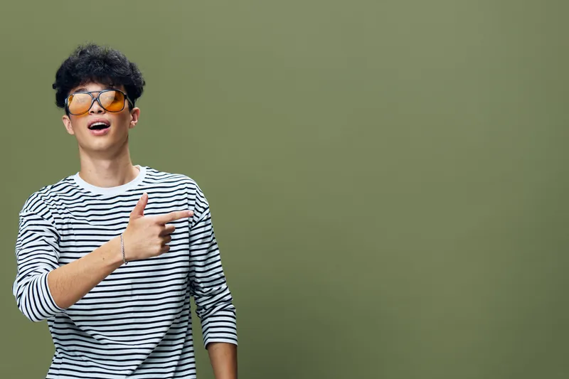 young man wearing sunglasses and striped shirt smiling and pointing with enthusiasm against olive green background, casual and trendy style portrait