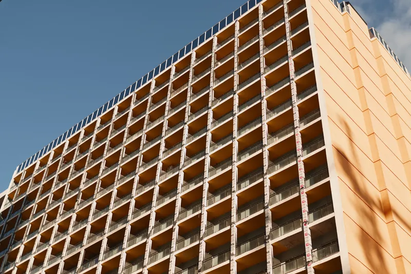 A high rise building under construction with an orange facade and grid balconies rises against a clear blue sky, showcasing industrial architecture and daytime activity.