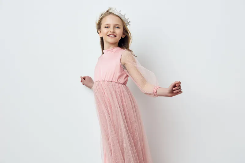 A joyful young girl in a pink dress with a delicate tulle skirt, smiling against a minimalist white background, showcasing happiness and childhood joy