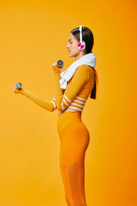Female athlete in bright orange activewear lifts dumbbells in a bold studio, wearing headphones and a towel around her neck, showcasing strength, focus, and energetic fitness training in a vivid
