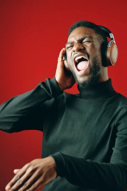 A young Black man passionately singing while wearing stylish headphones against a vibrant red background, showcasing joy and musical expression