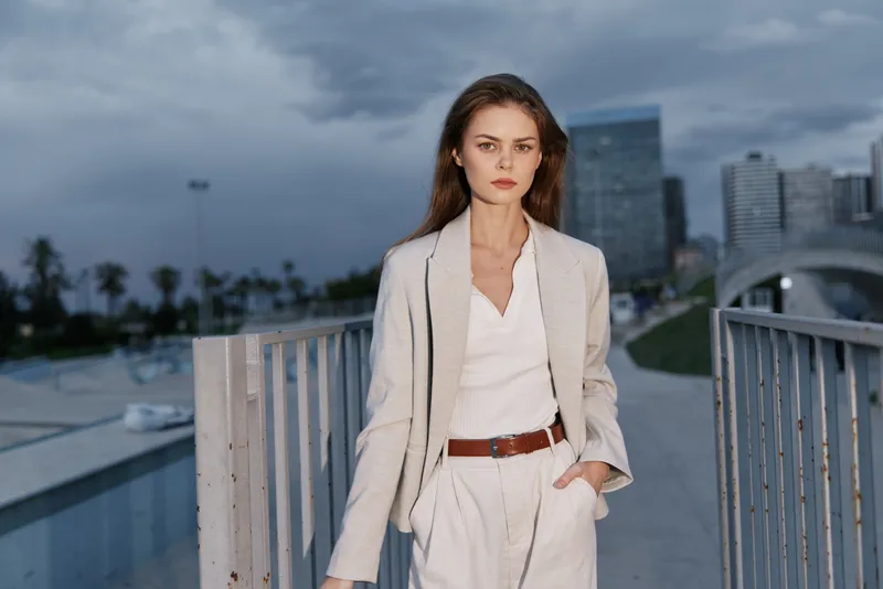 Young woman in beige business suit standing outdoors with city skyline and cloudy sky, confident and serious expression, urban fashion style