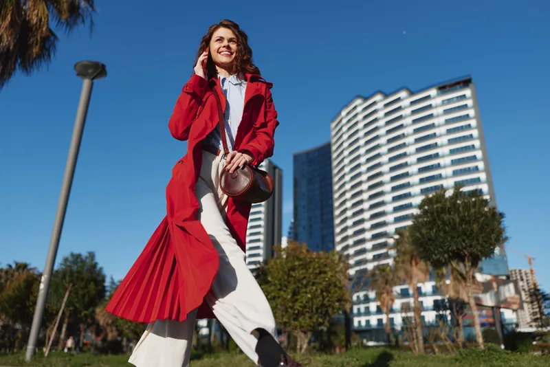 Cheerful woman outdoors, walking in a city park, red coat fluttering, white dress, sunny day, energetic pose, modern skyline, vibrant colors, candid moment