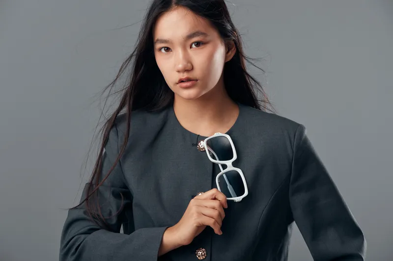 Portrait of confident young woman in dark blazer holding white sunglasses with long hair loose on gray background for fashion style and modern look.