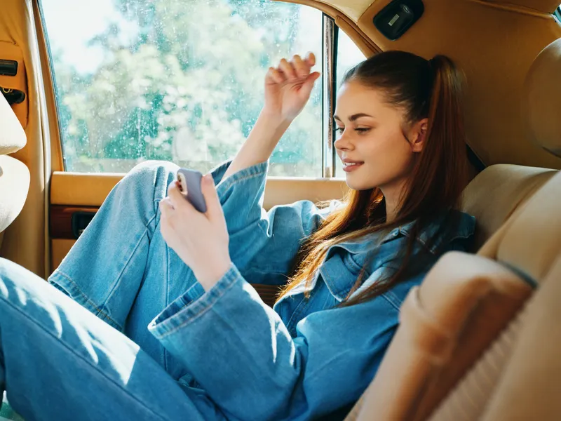 Young woman using smartphone in a vintage car with a relaxed expression, wearing denim outfit, enjoying a sunny day. Perfect for lifestyle and fashion concepts.