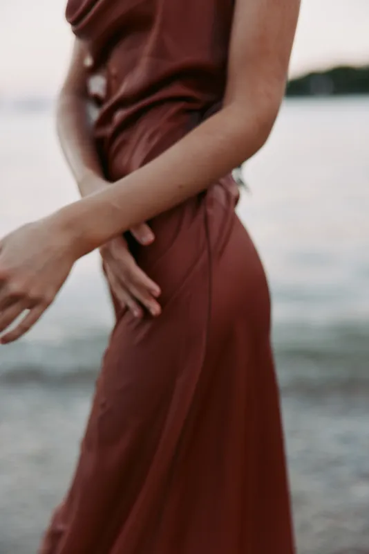 Close crop of a person in a rust red dress by a calm shoreline, the fabric draping softly as hands rest at the waist, creating a serene, intimate seaside moment.