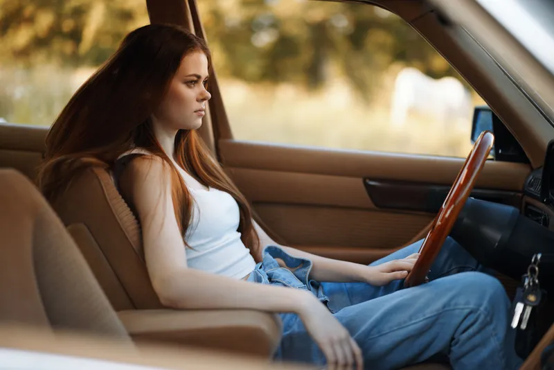 Young woman with long hair sitting in a vintage car, reflecting on life, dressed casually in a tank top and jeans, with a serene expression against a warm background.