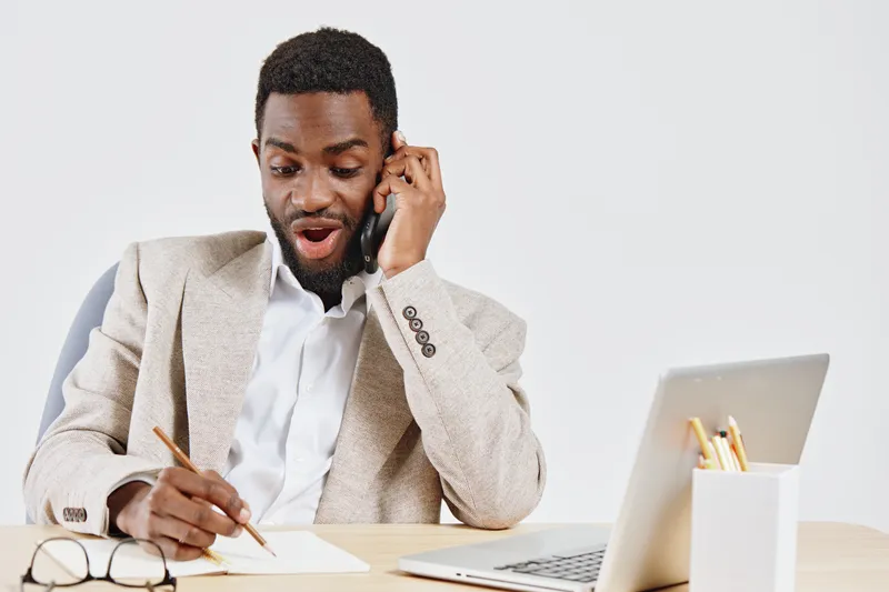 Professional young African American man in beige blazer and white shirt working at desk with laptop and notebook. Expressing surprise while talking on phone, on plain white background. Business commun