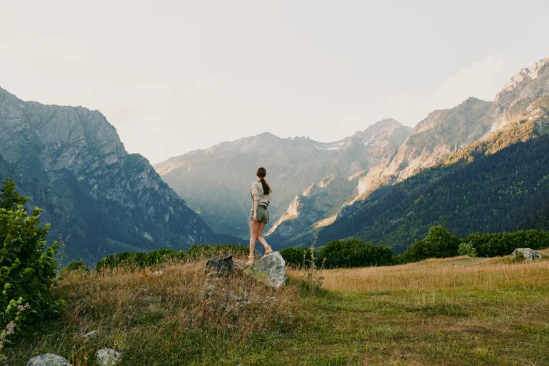Young woman stands on a grassy hillside with distant mountains rising, wearing a light summer dress, enjoying a peaceful outdoor moment as a gentle breeze moves through wildflowers under a clear sky.