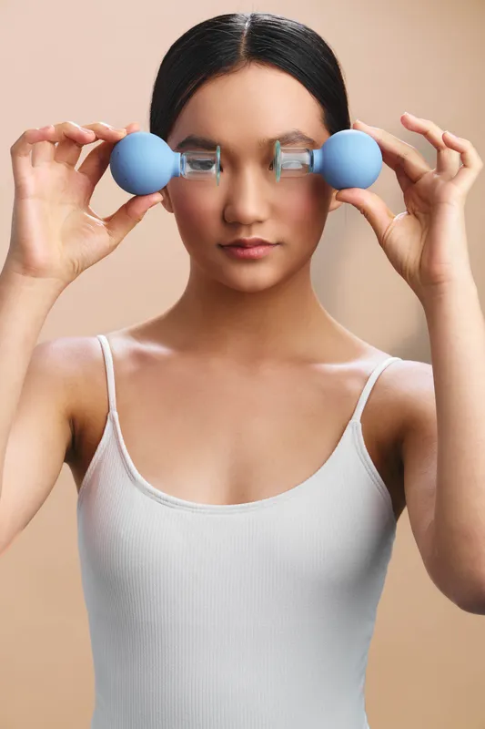 Asian woman performs a focused workout in a simple studio, holding blue dumbbells at eye level. She maintains balance, strength, and determined calm, showcasing healthy fitness.