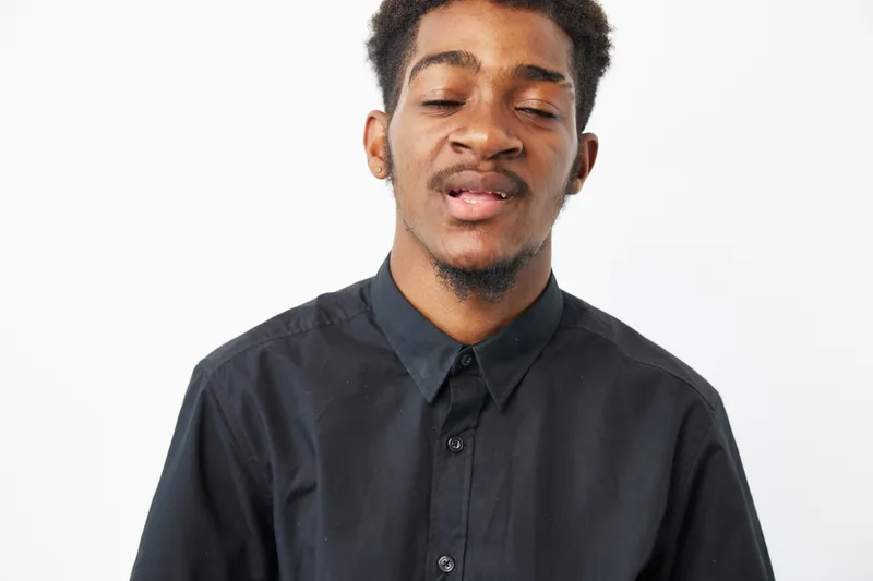 A young African American man with a playful expression, wearing a black shirt against a white background, exuding a sense of warmth and charm.