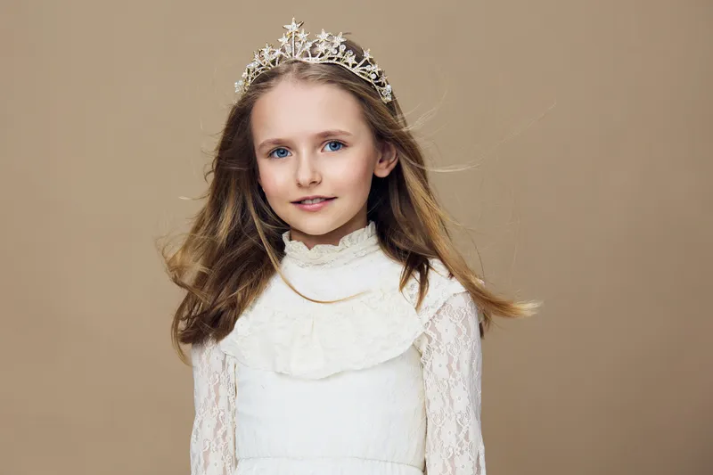 Young girl in a white dress and tiara against a beige background, conveying innocence and joy, perfect for children s fashion or fairy tale themes
