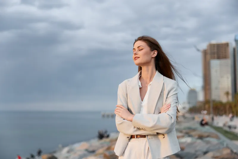 confident woman in light suit standing outdoors by sea coast with cityscape background during cloudy weather