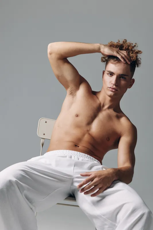Young man with curly hair posing shirtless sitting on chair with hand on head wearing white pants against gray background in studio. Male model with confident expression and casual posture.