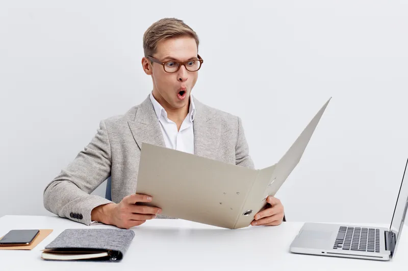Businessman looking surprised while reading document in office. Male professional in formal suit using laptop and paper sheet for work or presentation, studio shot with neutral background. People life