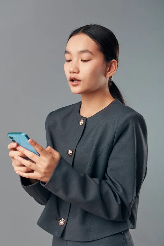 Young woman wearing gray formal jacket using smartphone with focused expression, standing against plain background for communication or technology concept in modern lifestyle.