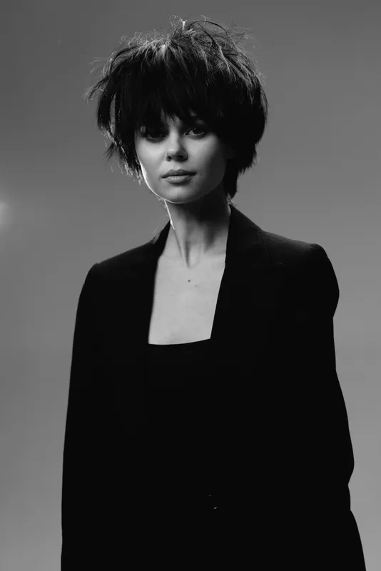 Black and white portrait of a young woman with short hairstyle wearing a dark blazer, expressionless with soft lighting and studio background.